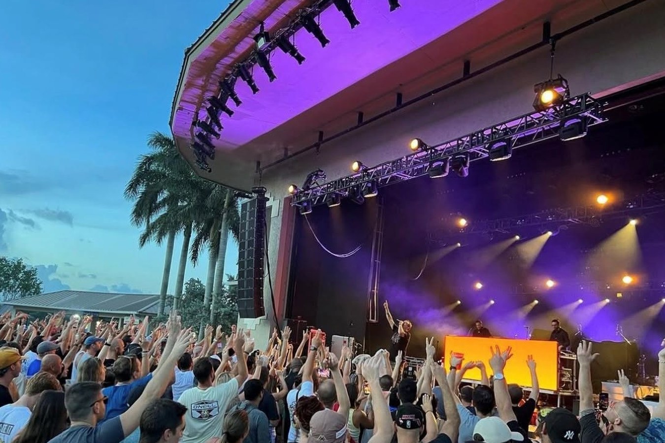 Crowd close to the Sunset Cove stage at dusk, hands in the air under purple and gold lights
